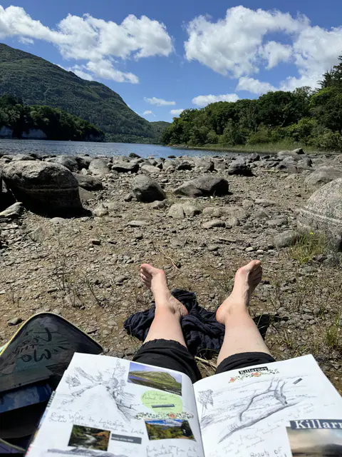 Pieds nus posés sur un rocher au bord d'un lac de montagne, un carnet de dessin ouvert sur les genoux.